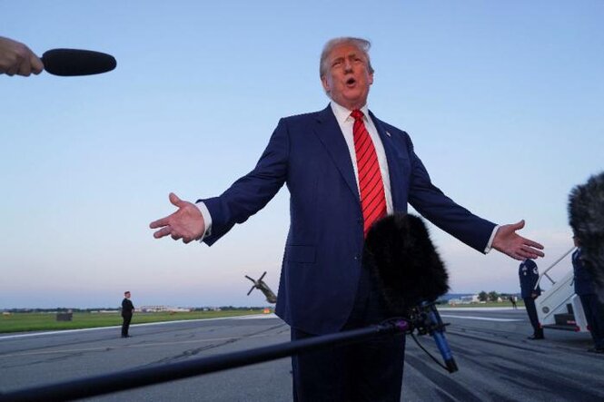 US-Präsident Trump steht auf einem Rollfeld mit ausgebreiteten Armen und spricht vor mehreren Mikrofonen. Im Hintergrund ist ein Flugzeug und das Rollfeld zu sehen.
