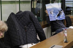 Zwei Männer sitzen auf der Anklagebank vor Gericht und verdecken ihre Gesichter mit einer Jacke und einem Poster