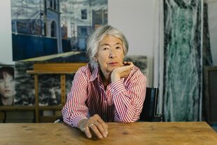 Evelyn Kuwertz sitzt am Tisch hinter ihr sind mehrere Malereien zu sehen