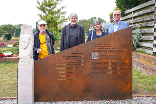 Denkmal für die Opfer der Kinderverschickung: Eine helle Stele mit einem weinenden Gesicht, an die sich eine Bronzetafel lehnt. Hinter der stehen drei Männer und eine Frau