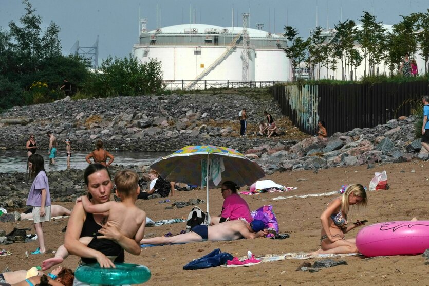 Menschen sonnen sich an einem Strand, im Hintergrund stehen große weiße Öltanks