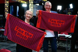 Ines schwerdtner und Jan van Aken halten rote Handtücher mit dem Spruch "tax the Rich" und dem Parteilogo in ihren Händen