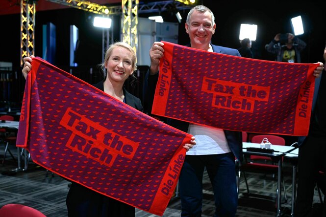 Ines schwerdtner und Jan van Aken halten rote Handtücher mit dem Spruch "tax the Rich" und dem Parteilogo in ihren Händen