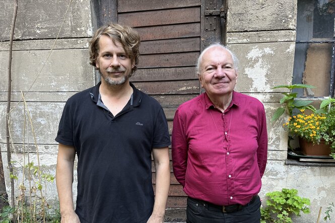 Die Musiker Kalle Kalima (links im Bild) und Conny Bauer (rechts im Bild) stehen im Freien vor einer unverputzen Hauswand und lächeln in die Kamera. Kalima trägt ein schwarzes T-Shirt, Bauer ein weinrotes Hemd.