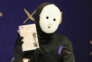 Uketsu mit weißer Maske vor dem Gesicht hält ein Buch in die Kamera