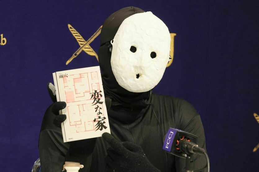 Uketsu mit weißer Maske vor dem Gesicht hält ein Buch in die Kamera