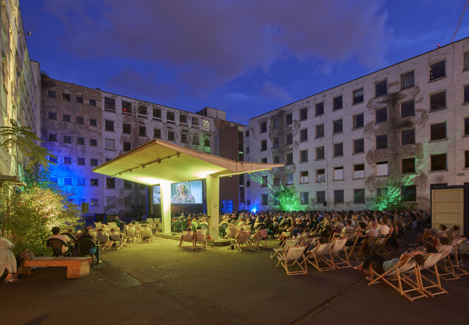 Menschen sitzen bei Einbruch der Dunkelheit auf Liegestühlen vor einer Leinwand im Freiluftkino Frankfurt, das im Innenhof des alten Polizeipräsidiums stattfindet.
