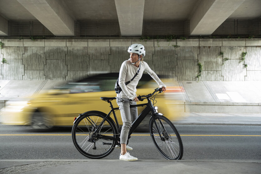 Eine Radfahrerin steht mit ihrem E-Bike am Bordstein, ein Auto fährt vorbei