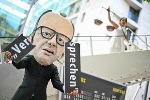 Merz-Figur mit einem zerbrochenen Schild, auf dem "Versprechen" steht
