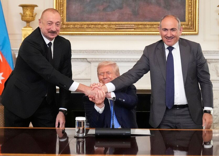 Aserbaidschans Präsident Ilham Aliyev (l) reicht Armeniens Ministerpräsident Nikol Paschinjan im State Dining Room des Weißen Hauses die Hand. Beide schauen lächelnd in die Kamera. Dahinter sitzt Donald Trump, grinst und hat eine Hand auf die Hände der beiden Männer draufgelegt