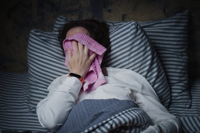Eine Frau im Bett presst sich einen rosa Waschlappen auf das Gesicht.