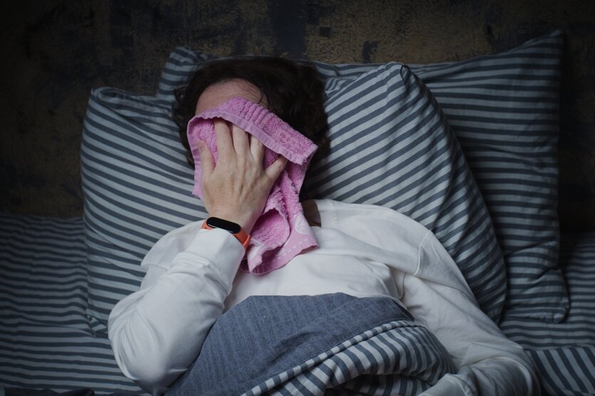 Eine Frau im Bett presst sich einen rosa Waschlappen auf das Gesicht.