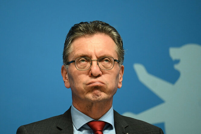 Gäbler bläst die Backen – Porträtfoto von Christian Gaebler, Berlins Senator für Stadtentwicklung, sitzt auch einer Pressekonferenz des Senats und schaut mit skeptischem Blick und leicht aufgeblähten Wangen nach oben.