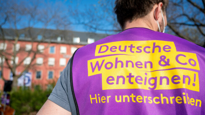 Eine Person von hinten, die eine lila Weste trägt. Auf der Weste steht in großer gelber Schrift: „Deutsche Wohnen & Co enteignen! Hier unterschreiben“. Im unscharfen Hintergrund ist ein Wohngebäude zu sehen.