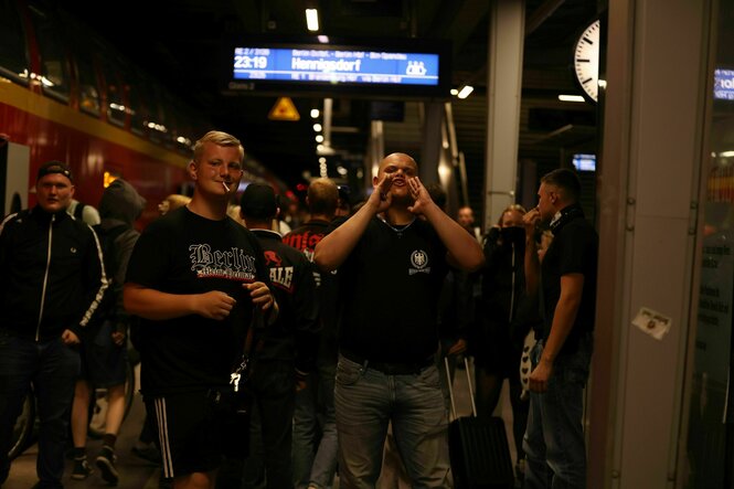 Männer in rechtsextremer Szenekleidung stehen an einem Bahnsteig und rufen etwas