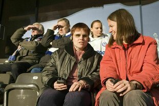 Sehbehinderte auf der Tribüne. Eine Frau trägt Kopfhörer, eine andere Frau in roter Jacke sitzt neben ihr und lacht
