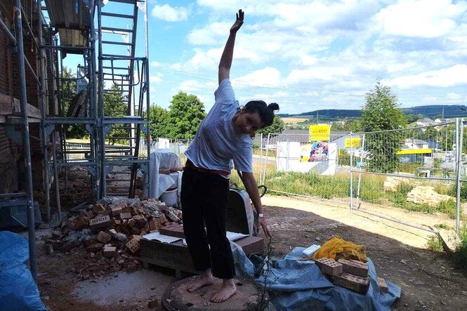 Eine junge Frau balanciert auf einem freistehenden Gullideckel vor einem Haus, das von einem Gerüst umstellt ist