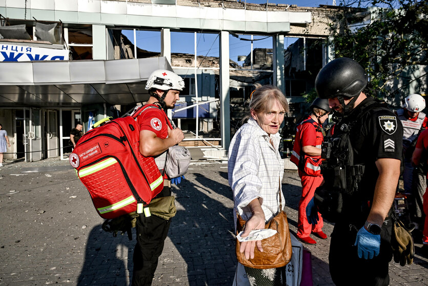 Ein zerbombtes Gebäude im Hintergrund, ein Polizist und ein Sanitäter kümmern sich um eine ältere Frau, die sehr erschrocken wirkt