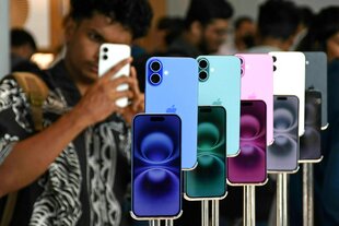 Smartphones von Apple in verschiedenen Farben stehen aufgereiht an einem Verkaufstich, davor stehen vor allem junge Männer, einer hält ein IPhone in der Hand