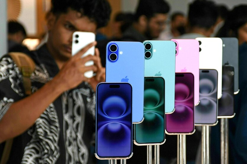 Smartphones von Apple in verschiedenen Farben stehen aufgereiht an einem Verkaufstich, davor stehen vor allem junge Männer, einer hält ein IPhone in der Hand