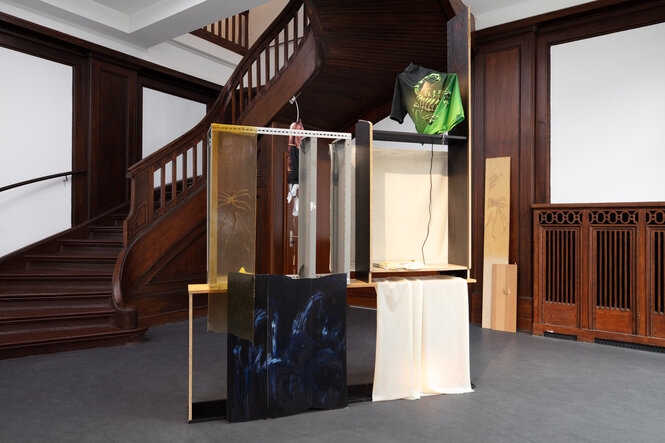 Blick in das Foyer des Künstlerhaus Bethanien. In der Mitte des Raumes steht eine Rauminstallation aus Billy-Regalen und verschiedenen Werkstoffen von Krys Huba. Oben an der Installation ist ein grünes T-Shirt befestigt, auf dem ein Skorpion zu sehen ist.