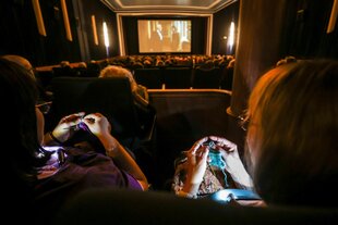 Im Vordergrund, über die Schulter, durch eine Sitzreihe gesehen, stricken zwei Frauen, im Mittelgrund ein Kinosaal mit menschen und abgeschlossen von einer Leinwand mit Film