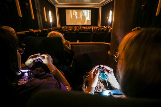 Im Vordergrund, über die Schulter, durch eine Sitzreihe gesehen, stricken zwei Frauen, im Mittelgrund ein Kinosaal mit menschen und abgeschlossen von einer Leinwand mit Film
