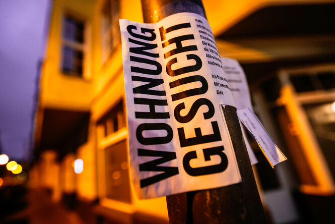 Nahaufnahme eines Zettels, der bei Nacht um einen Laternenpfahl oder einen Pfosten gewickelt ist. In großer, fetter, schwarzer Schrift steht darauf "WOHNUNG GESUCHT". Die Szene wird von warmem, künstlichem Licht beleuchtet, der städtische Hintergrund mit einem Gebäude und weiteren Lichtern ist unscharf.
