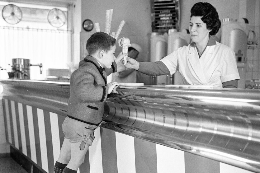 Ein historisches Foto von einem Kind, das sich über eine Theke beugt und eine Person hinter der Theke, die ihm eine Kugel Eis in der Waffel anreicht