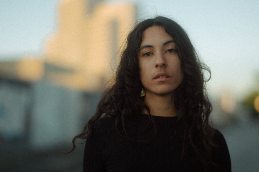 Die Musikerin Marie Tjong Ayong steht im Freien auf einer Straße. Hinter ihr deuten sich Hochhäuser an. Sie trägt einen schwarzen Pullover und lange Haare.