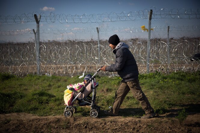 Das Bild zeigt einen warmgekleideten Menschen, der entlang eines Stacheldrahtzauns läuft und einen Kinderwagen schiebt, in dem ein Kind sitzt.