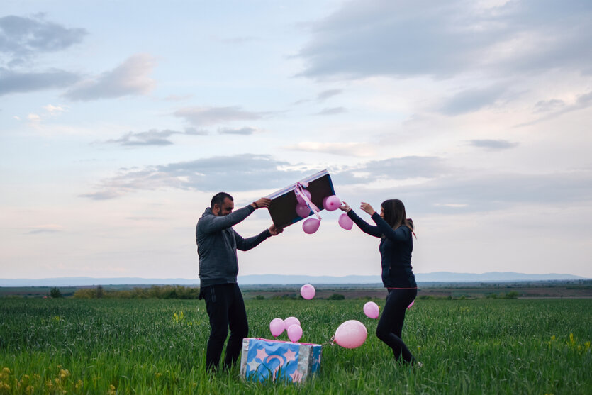 ein Mann und eine Frau auf einer Wiese öffnen eine Box aus der pinke Ballons aufsteigen