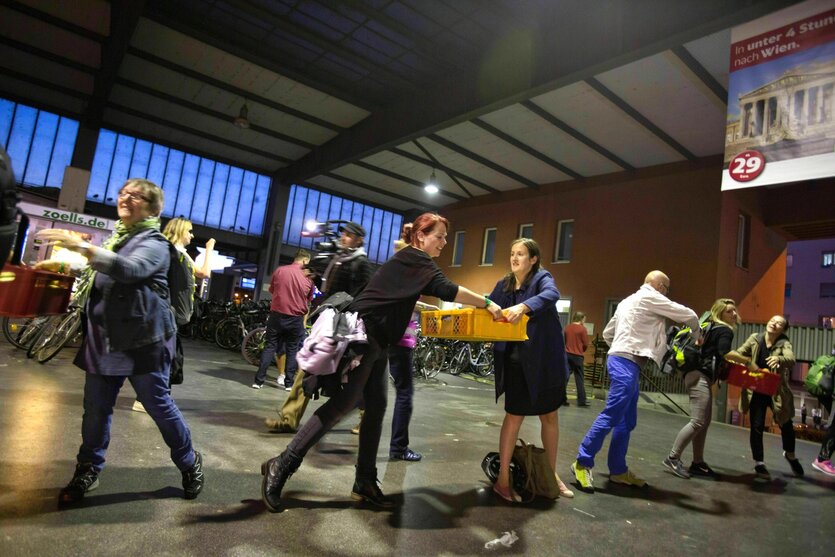 Menschen bilden im Bahnhof München eine Kette, um Kisten mit Lebensmitteln weiter zu reichen, dabei haben sie gute Laune