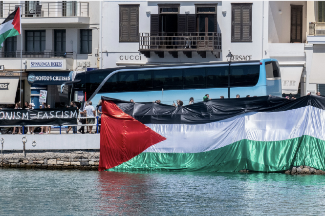 Zu sehen ist der Zugang zu einem Kreuzfahrtschiff in Griechenland. Der gesamte Zugangsbereich wird von einer Palästina-Fahne verdeckt, die von mehreren Menschen gehalten wird, während sie das Schiff betreten. Daneben, leicht abgeschnitten, erkennt man einen Teil eines Schilds, auf dem "Zionism Fascism" steht.
