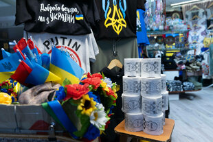 In einem Kaufhaus stehen verschiedene Produkte zum Verkauf: u.a. kleine ukrainische Flaggen, Plastikblumen und Toilettenpapier mit dem Gesicht vom russischen Präsidenten Putin dekoriert