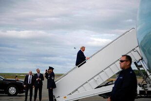 Trump steigt eine Gangway zur Präsidentenmaschine hoch. Am Fuß der Gangway stehen mehrere Personen. Im Vordergrund ist ein Sonnebrille tragender Security-Mann zu sehen
