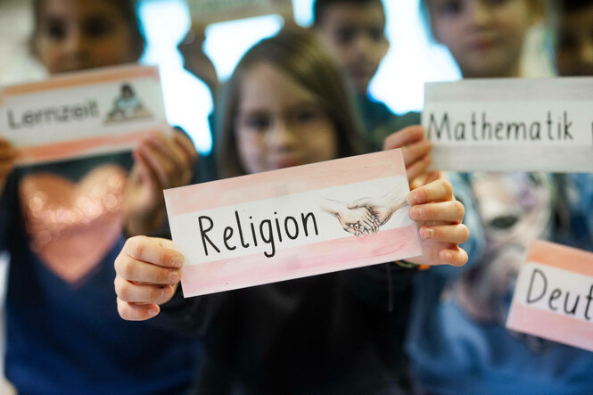 Schüler:innen halten Schilder auf denen Schulfächer, wie "Religion" oder "Mathematik" stehen