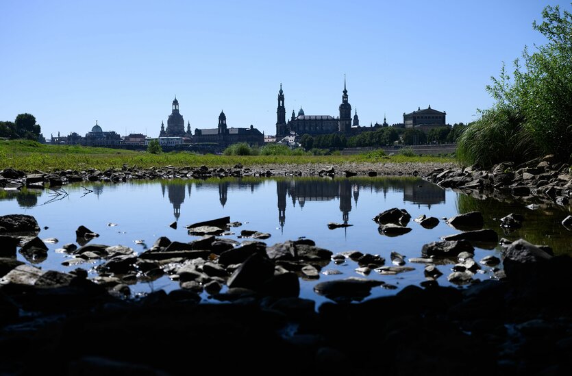 Die historische Altstadtkulisse mit der Kuppel der Kunstakademie (l-r) mit dem Engel „Fama“, der Frauenkirche, dem Ständehaus, der Hofkirche, dem Hausmannsturm und der Semperoper spiegelt sich unterhalb eines Brückenbogens der Marienbrücke in einer Wassersenke inmitten des ausgetrockneten Elbufers.