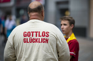 Ein Mann mit einem weißen Tshirt mit der Aufschrift "Gottlos Glücklich" steht mit dem Rücken zur Kamera