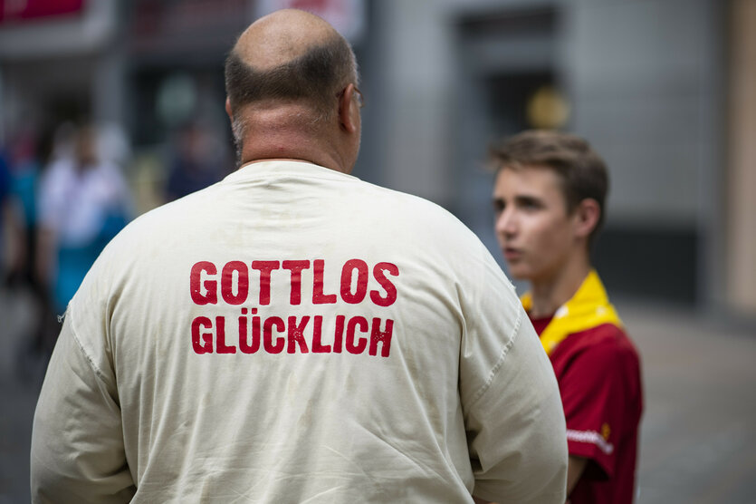 Ein Mann mit einem weißen Tshirt mit der Aufschrift "Gottlos Glücklich" steht mit dem Rücken zur Kamera