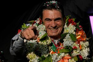 Ein Mann mit einem opulenten Blumenkranz um den Hals lacht und zeigt mit dem Finger auf sein Gegenüber
