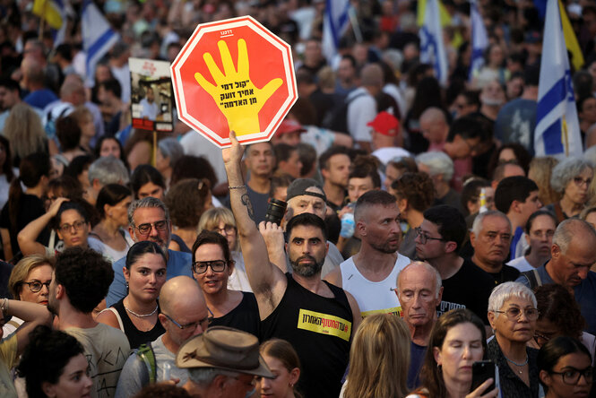 Ein Mann in einer Menschenmenge hält ein Plakat in Form eines Stop-Schildes darauf steht in hebräisch "Die Stimme des Blutes deines Bruders schreit aus der Erde"
