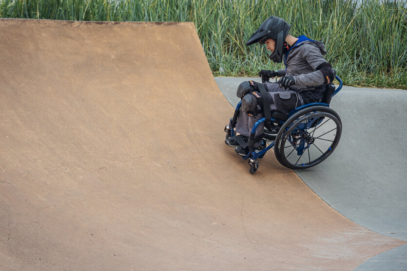 Rollstuhlfaher beim Skaten. Er trägt einen Helm und schaut konzentriert auf die Halfpipe.