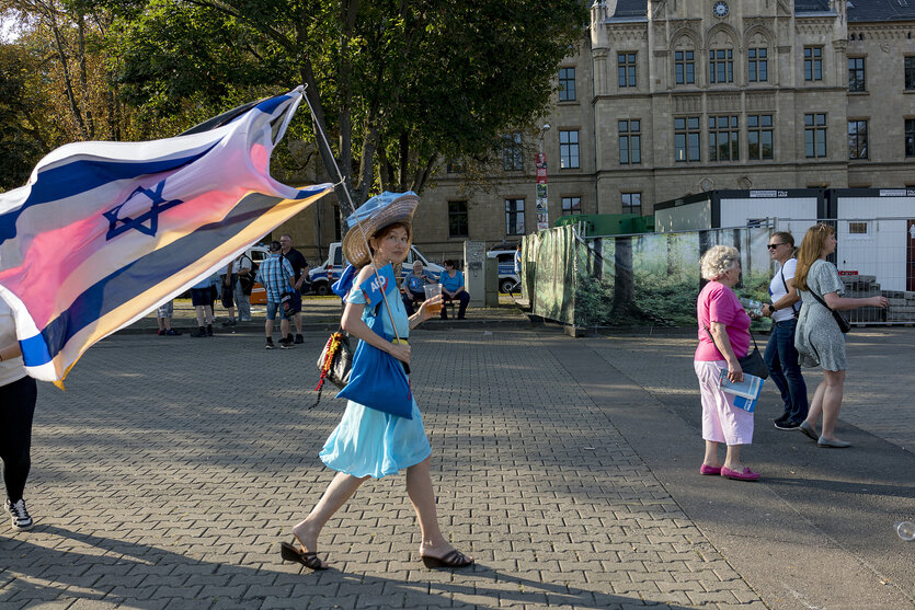Eine Frau im blauen Kleid trägt sowohl eine Israelfahne als auch ein Fähnchen mit dem Parteilogo der AfD
