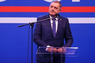 Mann mit Anzug und Krawatte hinter einem gläsernen Rednerpult