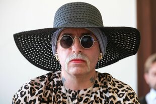 Marla-Svenja Liebich sitzt mit großem Hut, Sonnebrille und Schnurrbart im Gerichtssaal