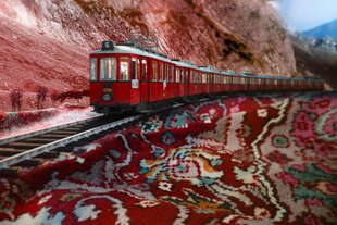 Ein roter Zug fährt auf einer Schienenstrecke durch eine bergige Landschaft. Die Umgebung ist von rötlich gefärbten Felsen und vereinzelten Bäumen geprägt. Ungewöhnlicherweise erscheint der Boden unter den Schienen wie ein buntes, orientalisches Muster, das an einen Teppich erinnert, mit roten, grünen und beigen Farbtönen. Der Himmel ist teilweise bewölkt, und die Szene hat einen surrealen, künstlichen Eindruck.