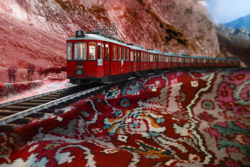 Ein roter Zug fährt auf einer Schienenstrecke durch eine bergige Landschaft. Die Umgebung ist von rötlich gefärbten Felsen und vereinzelten Bäumen geprägt. Ungewöhnlicherweise erscheint der Boden unter den Schienen wie ein buntes, orientalisches Muster, das an einen Teppich erinnert, mit roten, grünen und beigen Farbtönen. Der Himmel ist teilweise bewölkt, und die Szene hat einen surrealen, künstlichen Eindruck.