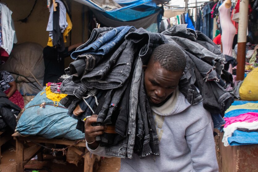 Ein Mann trägt einen Stapel Hosen zwischen Kleiderständen auf einem afrikanischen Markt