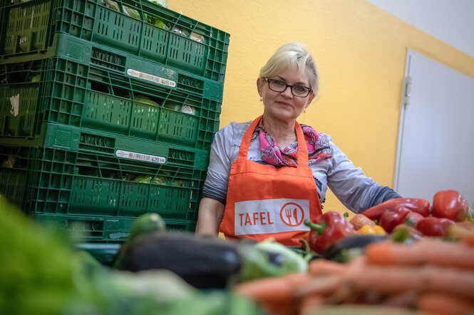 Eine Frau in orangefarbener Schürze mit dem Aufdruck "Tafel" steht hinter einem Berg Gemüse